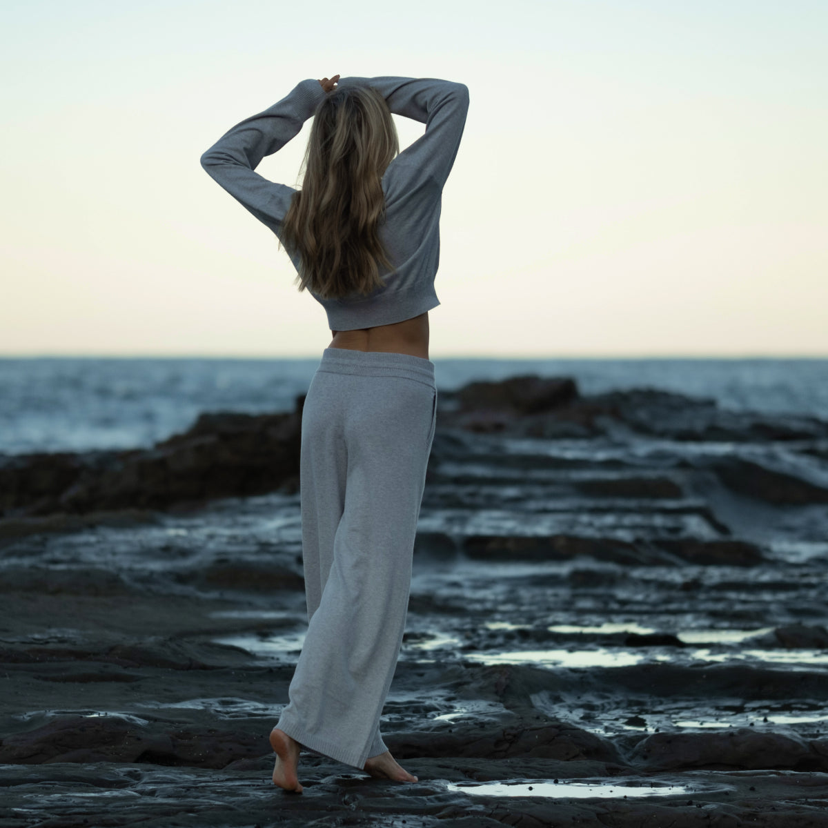 Model wearing grey knit jumper and wide-leg pants standing barefoot on rocky shore at sunset